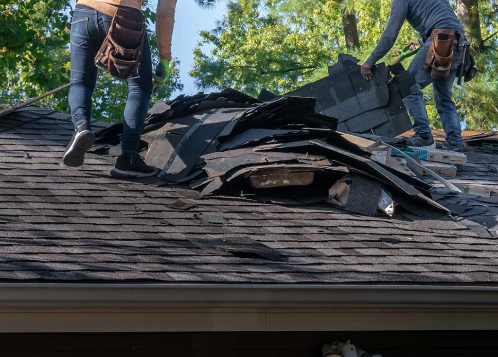 A roofing contractor inspects a shingled suburban home roof while holding a clipboard.